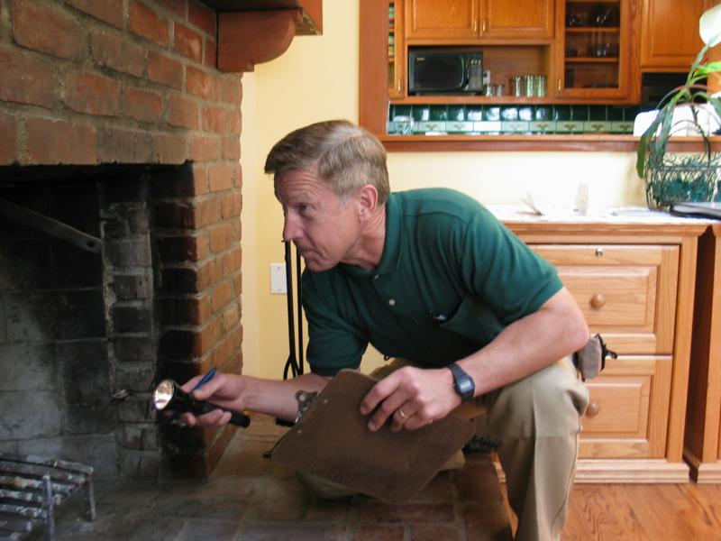 A technician kneeling at a brick fireplace with a flashlight and clipboard, inspecting it