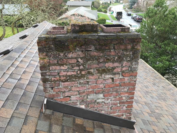 A weathered brick chimney on a shingled roof before repair